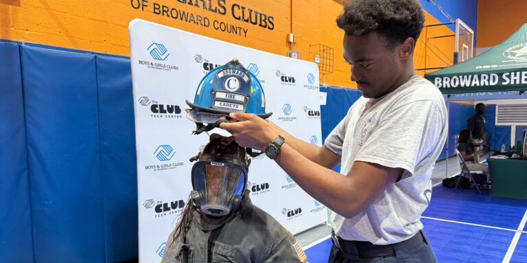 A boys and girls club member wearing a firefighter uniform, with a local firefighter placing a helmet on their head.