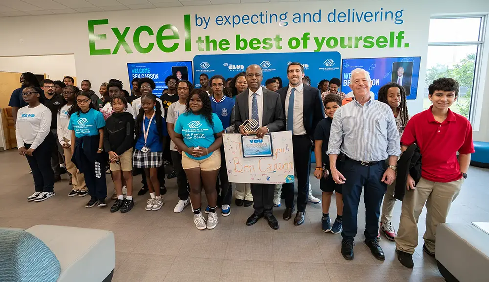 Dr. Ben Carson posing with BGCBC Club members and staff. Dr. Ben Carson is holding a handwritten sign that reads 'Thank you Dr. Ben Carson' and he holds a statue of the Boys & Girls Clubs of Broward County Logo.