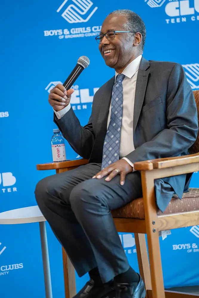 Dr. Den Carson smiling while holding a microphone. He sits in a chair in front of a BGCBC Branded back drop