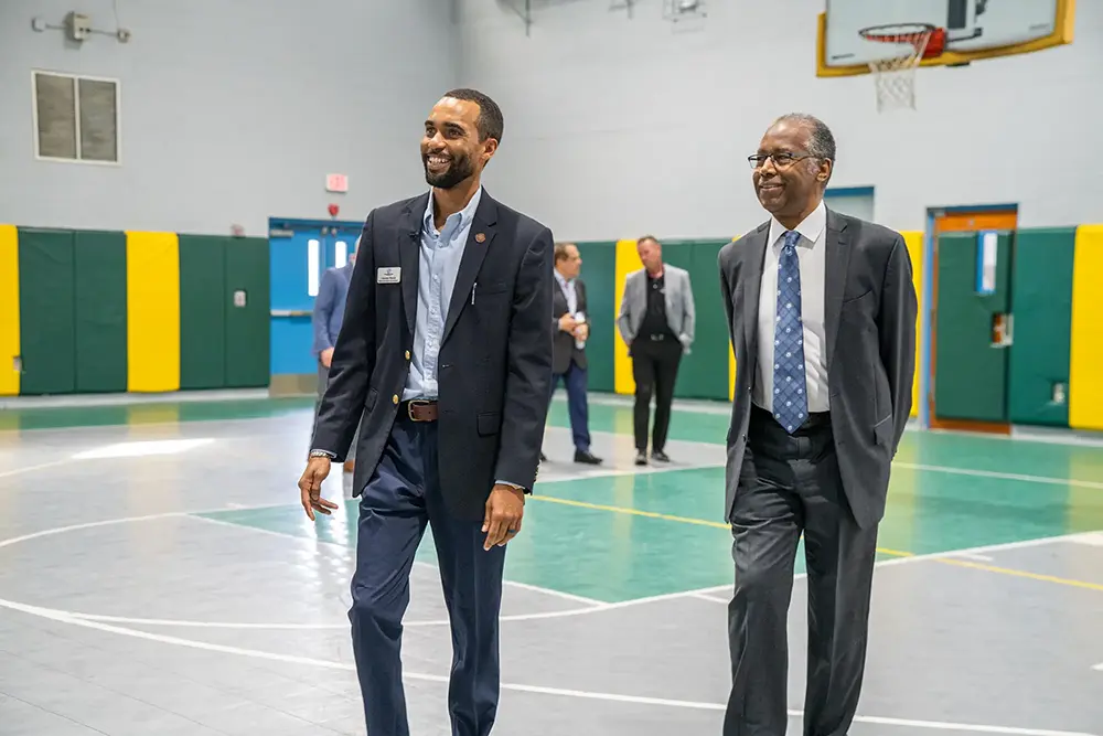 Dr. Ben Carson smiling as he walks with a BGCBC administrative staff member. They walk in the Teen Centers gym.