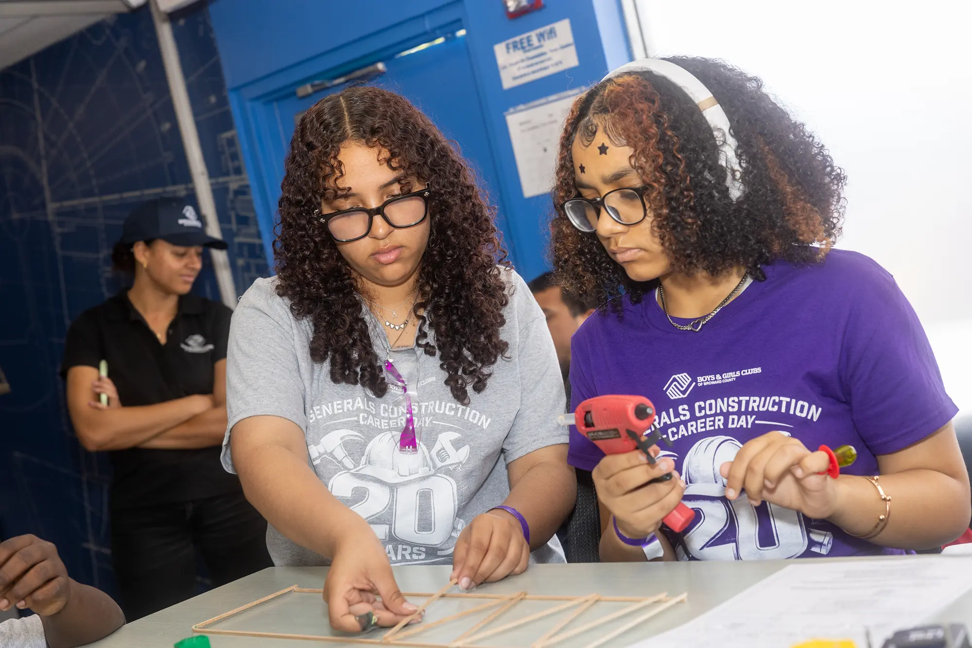 Two BGCBC Club members working together to hot glue popsicle sticks together. Both club members are wearing Generals Construction Career Day 20 Years' shirts.
