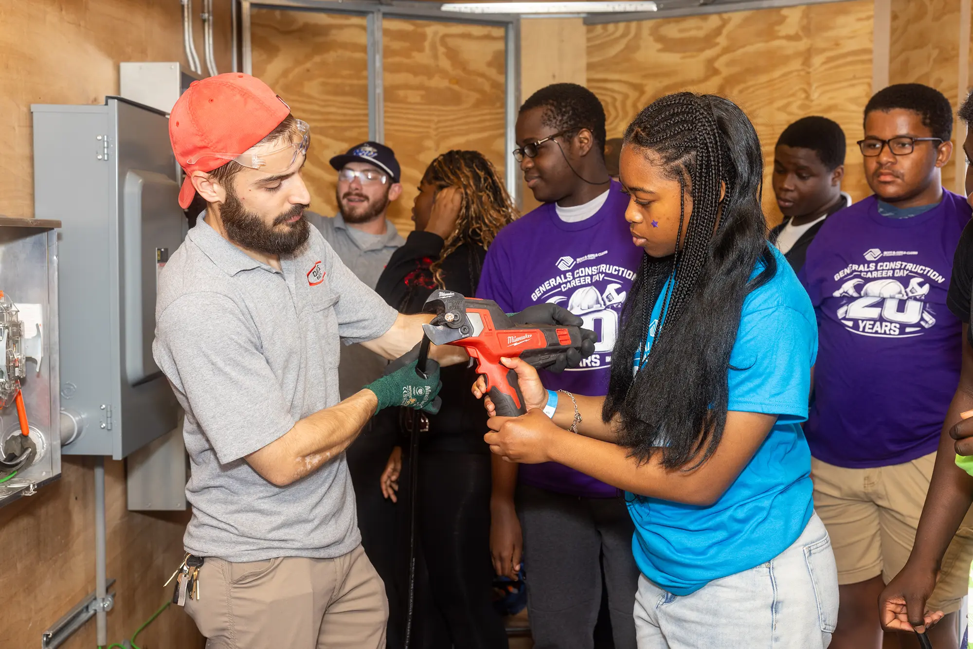 A Boys & Girls Clubs of Broward County member being guided by an industry professional while she cuts an electrical cord. BGCBC Club members watch behind her, wearing 'Generals Construction Career Day 20 years' shirts.