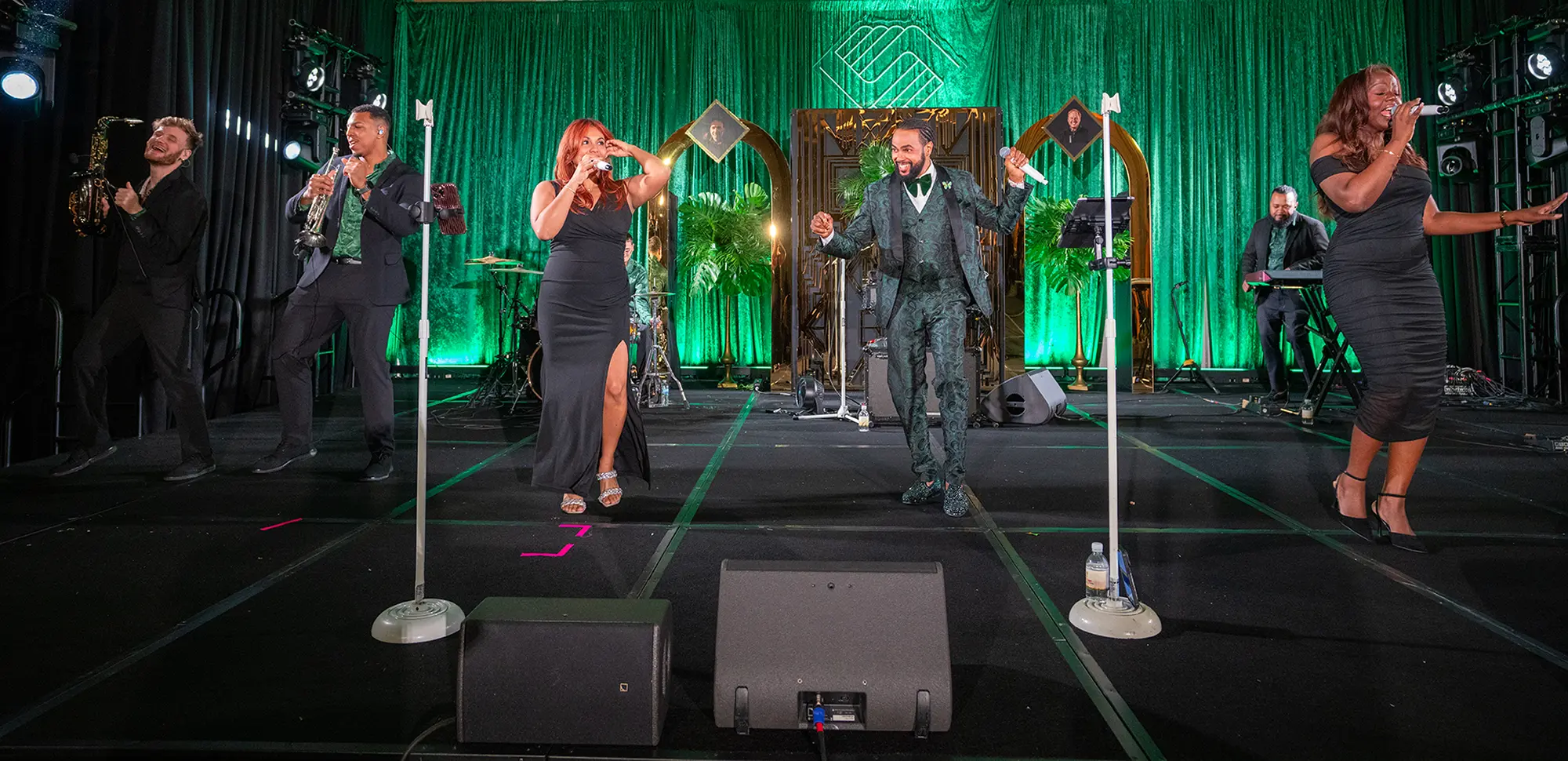 Live Music entertainment from the event. Of the five performers, three can be seen holding microphones, one holds a trumpet, and the remaining performer holds a saxophone. All five performers are dancing and smiling on stage.
