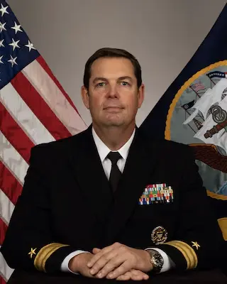 Rear Admiral Ryan Perry in his decorated admiral navy service dress blue uniform. He sits in front of the Flag of the United States and the flag of United States Navy