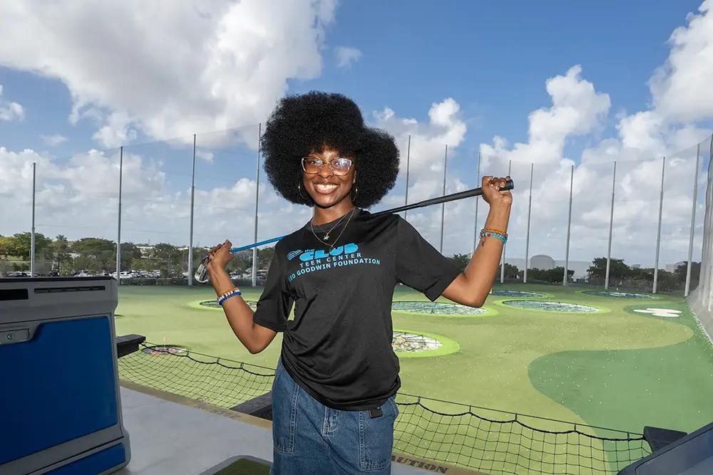 A Boys & Girls Clubs of Broward County Club member smiling holding a golf club on her shoulders. She wears a black 'The Club Teen Center' shirt.