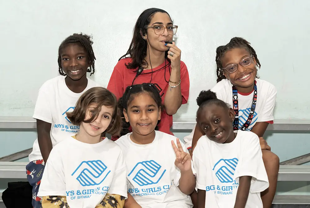 A BGCBC staff member blowing into a whistle with five BGCBC Club members posing alongside them. The club members each wear a white shirt with blue text that reads 'Boys & Girls Club of Broward County'