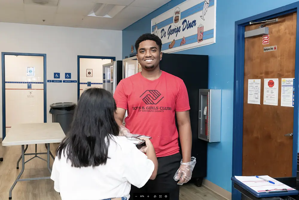 A BGCBC staff member wearing gloves as they serve an after school snack to a BGCBC Club member. The staff member is wearing a red shirt with black letter that reads 'Boys and Girls Clubs of Broward County.