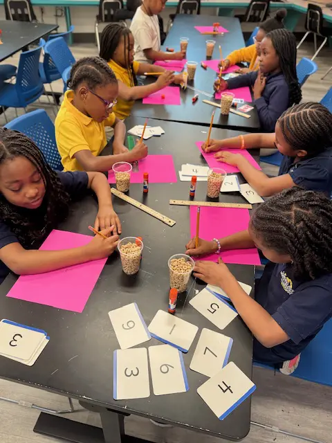 BGCBC Club members solving math problems on pink pieces of paper. Each club member has a cup of cheerios to help them count out the equation visually