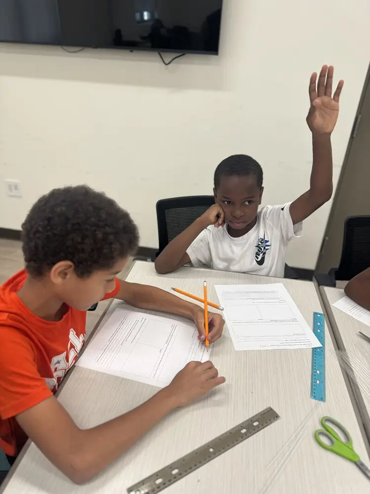 Two BGCBC club members sitting at a table. One club member is working on a work sheet while the other is raising their hand