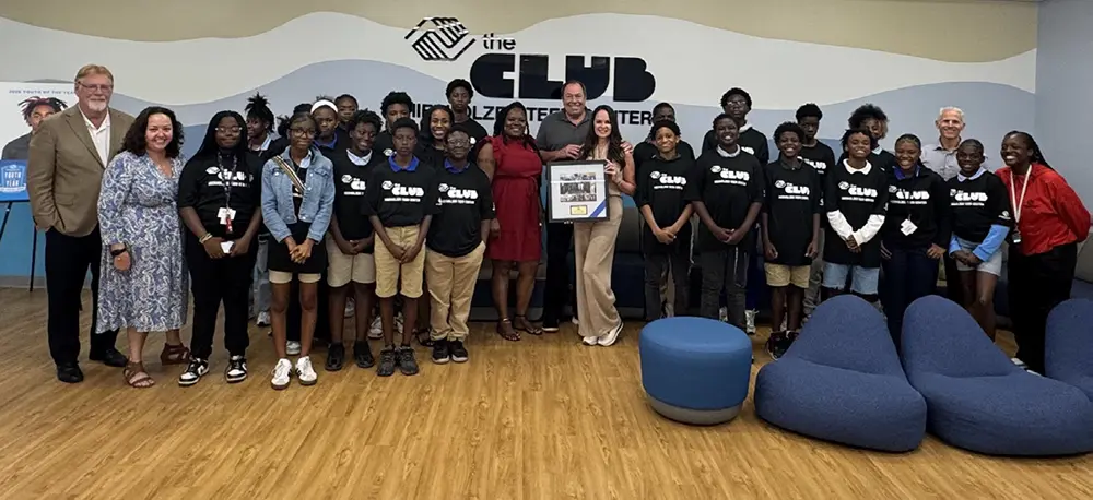 A group photo of Christy and Larry Hierhozler holding a framed image from the club ribbon cutting. Christy and Larry Hierhozler are posing with BGCBC Club members, staff, and administrative staff.