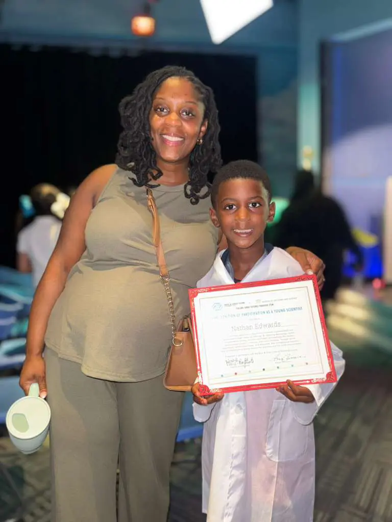 A BGCBC Club Member posing with their Mom at the Family Science Night. The club member is wearing a lab coat and holding the certificate they earned from the event.