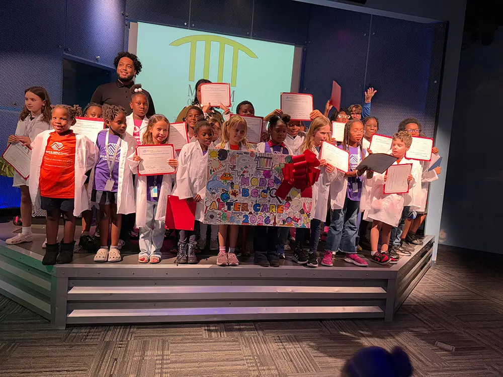 A group picture of the BGCBC Club members that participated in the Family Science Night. Each club member is holding a certificate and wearing a lab coat from the event.
