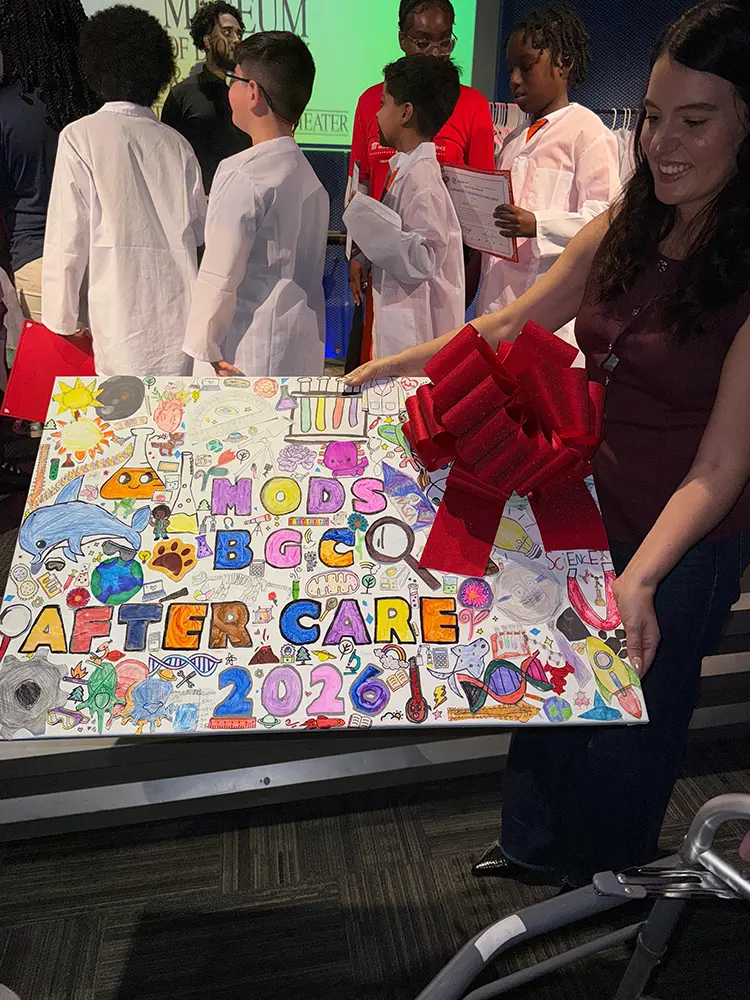 An administrative BGCBC Staff member holding a hand-drawn sign with Science imagery that reads "MODS BGC After Care 2026".